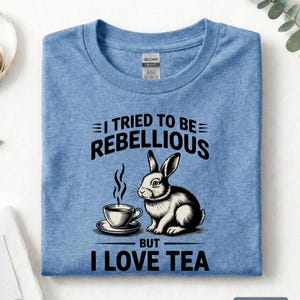 Puede incluir: Camiseta azul &iacute;ndigo jaspeado con el texto "I TRIED TO BE REBELLIOUS BUT I LOVE TEA" y una ilustraci&oacute;n en blanco y negro de un conejo y una taza de t&eacute;. La camiseta es de cuello redondo.