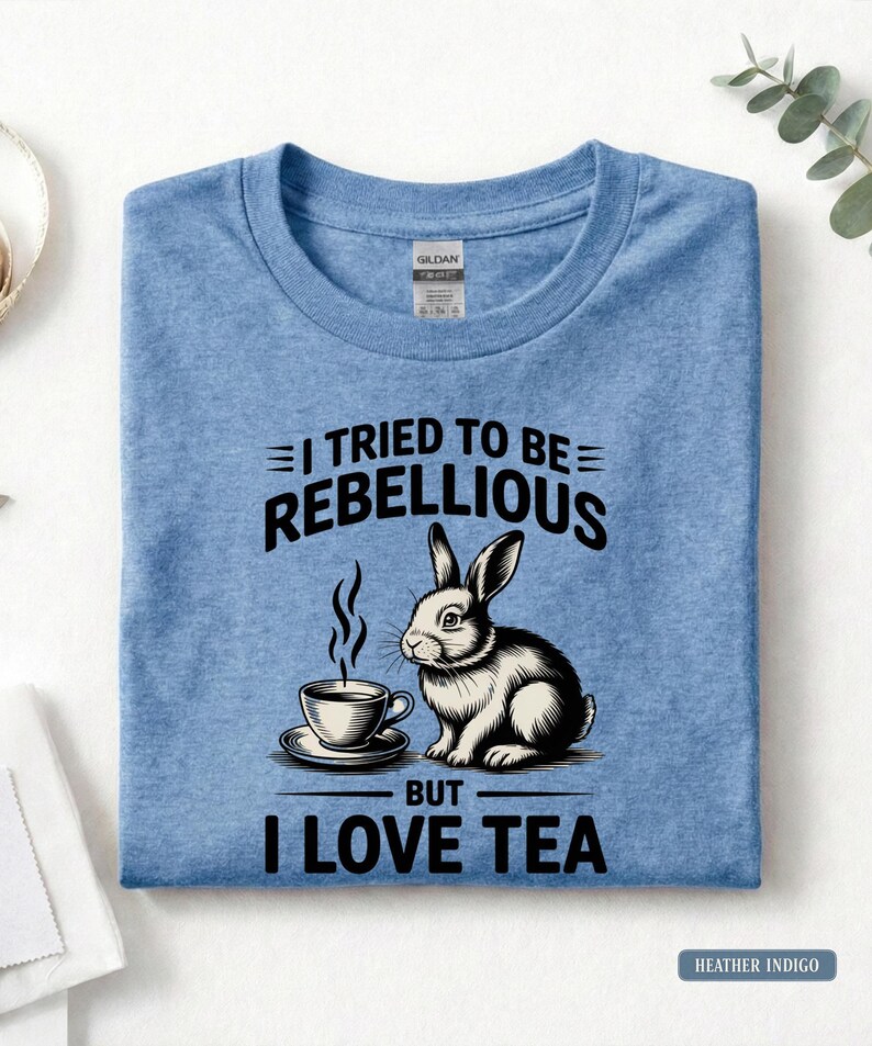 Puede incluir: Camiseta azul &iacute;ndigo jaspeado con el texto "I TRIED TO BE REBELLIOUS BUT I LOVE TEA" y una ilustraci&oacute;n en blanco y negro de un conejo y una taza de t&eacute;. La camiseta es de cuello redondo.