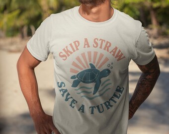 May include: A light beige t-shirt with the text "SKIP A STRAW SAVE A TURTLE" in a circular design. The design includes a turtle and sunburst graphic in blue and coral colors. The shirt is a crew neck.