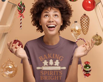 May include: A woman wearing a mauve t-shirt with the text "BAKING SPIRITS BRIGHT" in brown and cream. The image is festive, with Christmas ornaments and a ladder in the background. The woman is smiling and looking upwards.