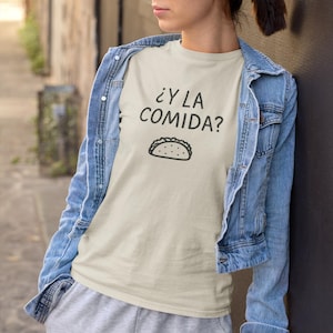 May include: A light beige t-shirt with the text "¿Y LA COMIDA?" and a taco illustration in black. The person is wearing a light blue denim jacket and gray sweatpants.