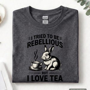Puede incluir: Camiseta gris oscuro con un dise&ntilde;o de un conejo y una taza de t&eacute;. El texto en negro dice: "I TRIED TO BE REBELLIOUS BUT I LOVE TEA". La camiseta est&aacute; doblada.