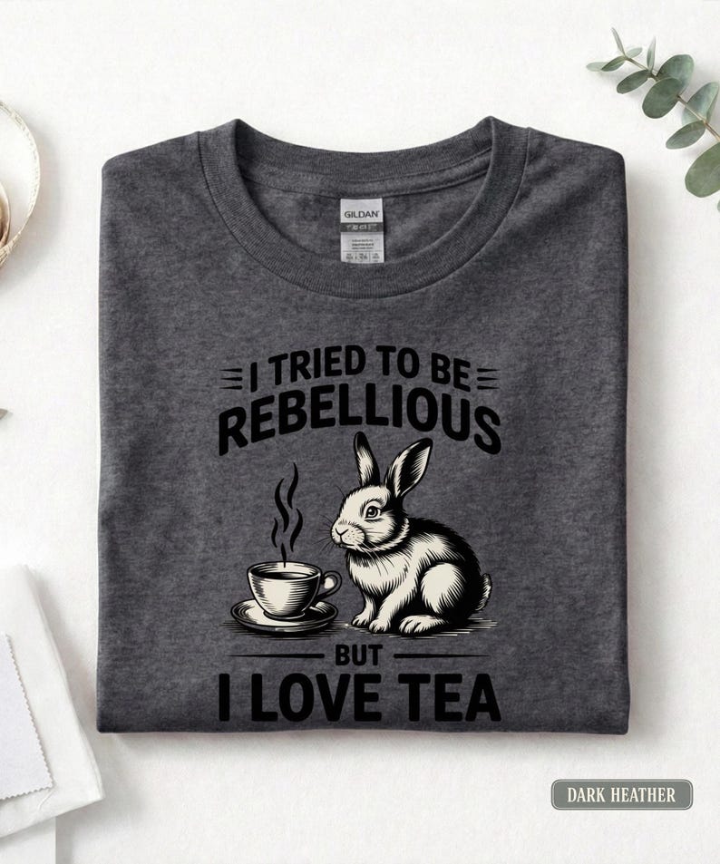 Puede incluir: Camiseta gris oscuro con un dise&ntilde;o de un conejo y una taza de t&eacute;. El texto en negro dice: "I TRIED TO BE REBELLIOUS BUT I LOVE TEA". La camiseta est&aacute; doblada.