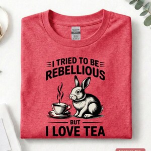 Puede incluir: Camiseta rojo jaspeado con un dise&ntilde;o de conejo y taza de t&eacute;. El texto dice "I TRIED TO BE REBELLIOUS BUT I LOVE TEA". La camiseta est&aacute; doblada.