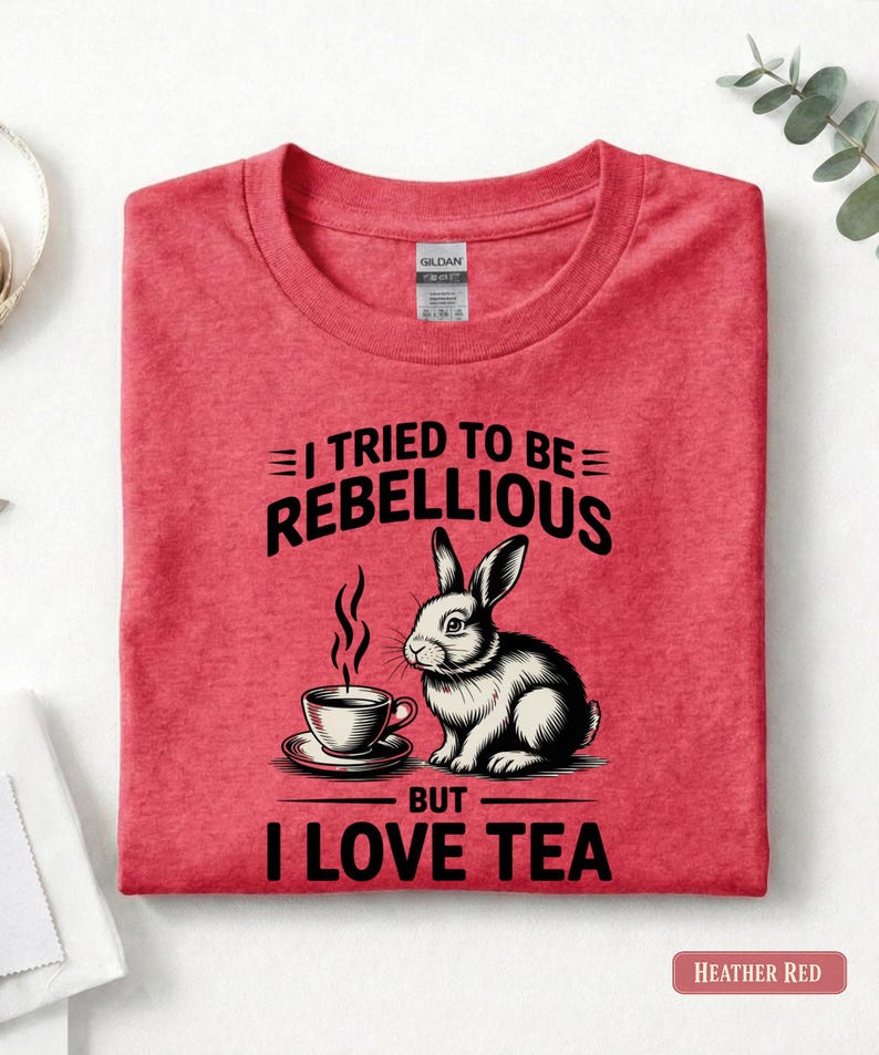 Puede incluir: Camiseta rojo jaspeado con un dise&ntilde;o de conejo y taza de t&eacute;. El texto dice "I TRIED TO BE REBELLIOUS BUT I LOVE TEA". La camiseta est&aacute; doblada.