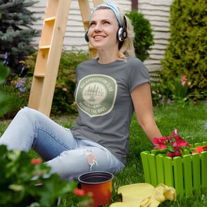 May include: A gray t-shirt with a circular graphic that reads "Bug Appreciation Club, Respect the Bugs." The shirt is worn by a person sitting on the grass, surrounded by flowers and gardening tools.