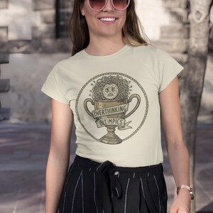 May include: Beige t-shirt with a humorous graphic of a trophy with a worried face and spaghetti-like hair, encircled by a rope design. The trophy has the words "Overthinking Olympics" on it.