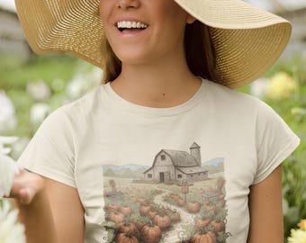 May include: A light beige t-shirt featuring a detailed illustration of a pumpkin patch with a barn in the background. The image shows a path leading through the pumpkins. The person is wearing a wide-brimmed straw hat.