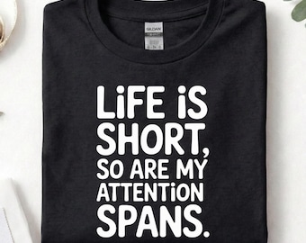 May include: Black t-shirt featuring the white text: "LIFE IS SHORT, SO ARE MY ATTENTION SPANS." The shirt is neatly folded. The word "BLACK" is printed at the bottom. A Gildan tag is visible.