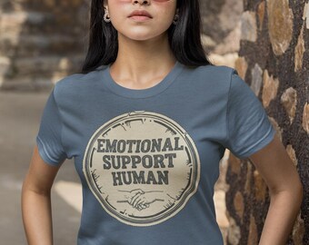 May include: A blue t-shirt with a circular graphic that says "EMOTIONAL SUPPORT HUMAN" in a vintage style. The graphic includes a handshake illustration. The t-shirt is plain and has short sleeves.
