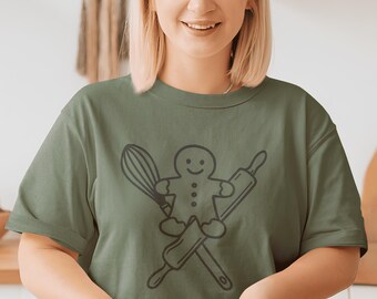 May include: Olive green t-shirt featuring a black graphic of a gingerbread man holding a whisk and rolling pin. The design is crossed over a rolling pin. The person is smiling and working with dough.