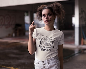 May include: A white t-shirt with a black graphic of a haunted house, trees, and the text "Grim Grinning Ghosts." The person in the image is wearing white jeans and has clown-like makeup.