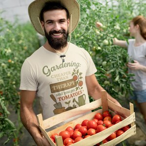 May include: A man wearing a straw hat and a t-shirt that says "Gardening: Cheaper Than Therapy and you get Tomatoes" holds a wooden crate filled with ripe red tomatoes. Another person is in the background, picking tomatoes in a greenhouse.