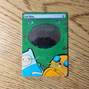 May include: A green card with the text "Sol Ring" and the number "1". The card features cartoon illustrations of Finn and Jake from Adventure Time. The background is green with a dark oval shape in the center.