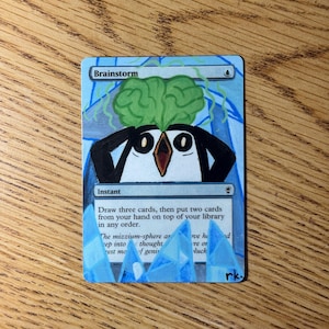 May include: A custom-painted "Brainstorm" Magic: The Gathering card. The card features a cartoon penguin with a green brain and blue accents. The text "Brainstorm" and card instructions are visible.