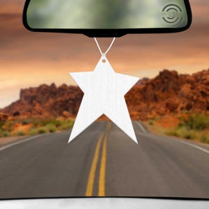 May include: A white star-shaped hanging ornament suspended by a thin white string. The star has a smooth, flat surface and five pointed tips. It is positioned centrally in the frame, hanging from a black rearview mirror against a desert road.