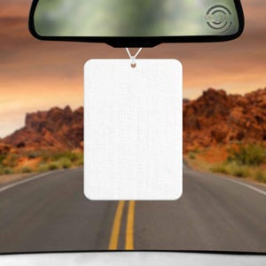 May include: A rectangular white car air freshener hanging from a rearview mirror by a thin white elastic string. The air freshener has a smooth, blank surface with rounded corners, suspended against a blurred background of a desert road at sunset.