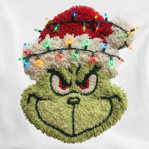 May include: White sweatshirt featuring a green Grinch face wearing a red and white Santa hat adorned with colorful Christmas lights. The Grinch's face has a textured, fuzzy appearance, with red eyes and a mischievous grin. A festive holiday-themed design.