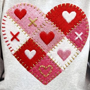 May include: A light gray sweatshirt featuring a large, patchwork heart design. The heart is composed of red, pink, and white squares, each adorned with heart shapes, crosses, and stitched details. A gold chain bracelet is visible on the wrist.