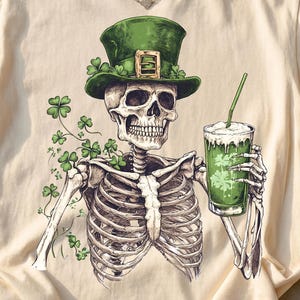 Op de afbeelding: Crèmekleurig T-shirt met een skelet met een groene St. Patrick's Day hoed en een groen drankje met een rietje. Het skelet is omringd door klavers. Het shirt heeft een ronde hals en korte mouwen.