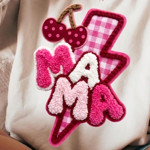 May include: Cream-colored sweatshirt with a pink and white gingham lightning bolt design. The lightning bolt has the word "MAMA" in a fluffy, textured font. Two cherries with brown stems are above the lightning bolt.