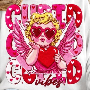 Puede incluir: Sudadera blanca con un gráfico de Cupido con gafas de sol en forma de corazón, sosteniendo un corazón y enviando un beso. Las palabras "Cupid Good Vibes" están en rosa y rojo, con acentos de corazón. Un top temático de San Valentín.