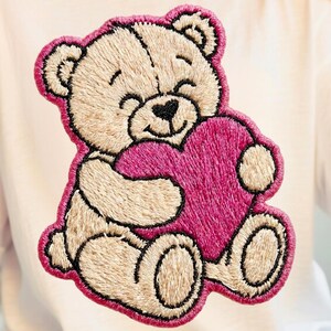 Könnte beinhalten: Weißes T-Shirt mit einem gestickten Teddybären, der ein rosa Herz hält. Der Teddybär hat ein lächelndes Gesicht und ist mit einem dunkleren Faden umrandet. Das Herz hat eine einfarbige rosa Farbe.