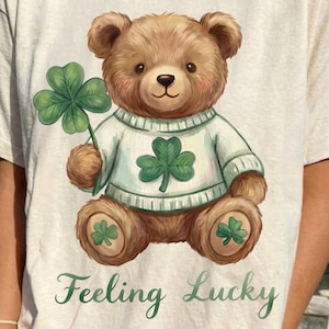 May include: Cream-colored t-shirt featuring a brown teddy bear holding a green four-leaf clover. The bear wears a white sweater with a green shamrock. The words "Feeling Lucky" are printed in green script below the bear.