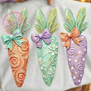 May include: White sweatshirt featuring three colorful carrot designs with bunny ears and bows. The carrots are orange, green, and purple, with floral and swirl patterns. The design is held by a person.