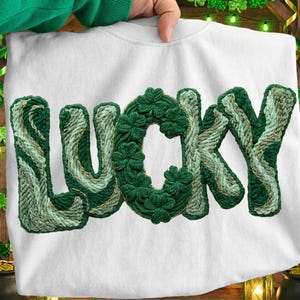 Könnte beinhalten: Weißes Sweatshirt mit dem Wort "LUCKY" in Grüntönen gestickt. Die Buchstaben haben eine strukturierte, gewebte Optik, wobei der Buchstabe "C" mit Kleeblatt-Designs gefüllt ist. Ein St. Patrick's Day-Thema.