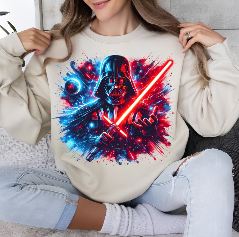 May include: A cream-colored sweatshirt with a graphic print of Darth Vader holding a red lightsaber. The background is a blue and red galaxy with stars and planets.