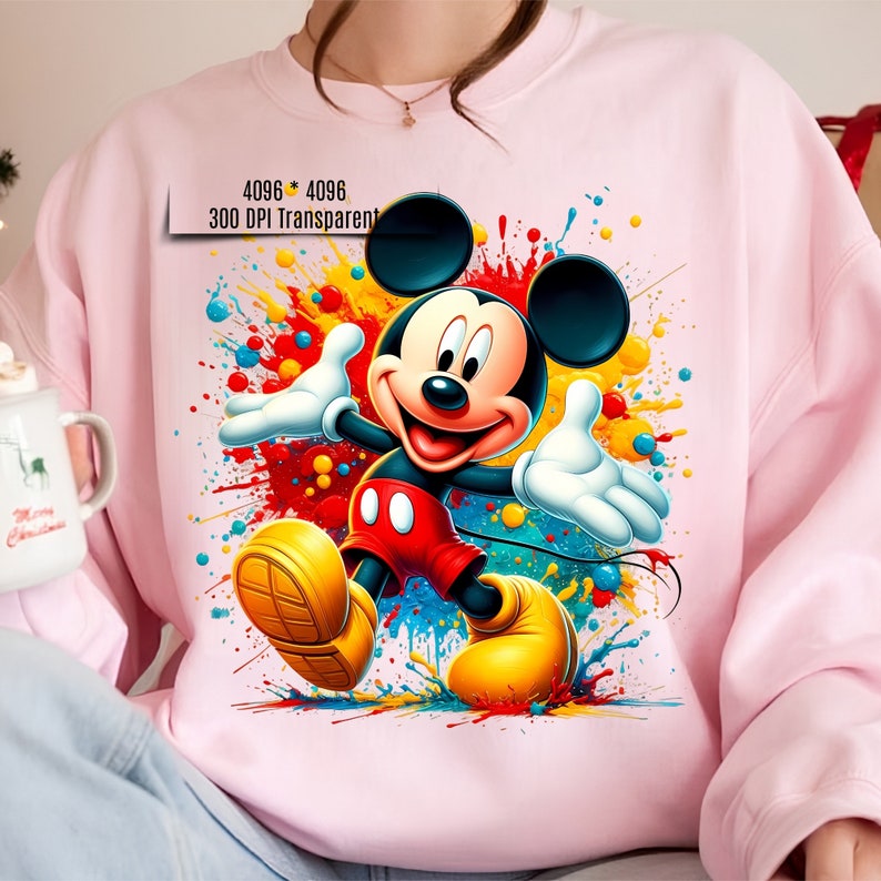 May include: Mickey Mouse cartoon character in a colorful paint splatter design. The character is wearing red shorts and yellow shoes. The background is a pink sweatshirt.