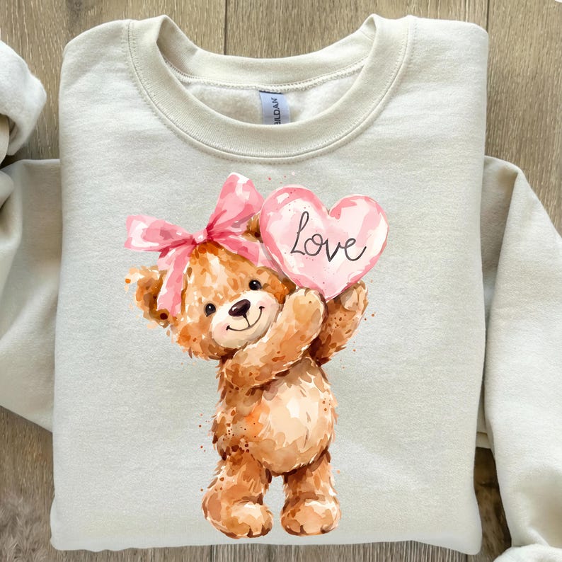 May include: A beige sweatshirt featuring a watercolor illustration of a teddy bear holding a pink heart-shaped sign with the word "Love" written on it. The teddy bear has a pink bow on its head.