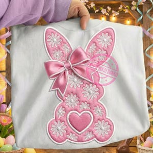 Könnte beinhalten: Weißes Sweatshirt mit einem rosa Hasen-Design. Der Hase ist mit weißen, gänseblümchenartigen Blumen und einer rosa Schleife verziert. Ein rosa Herz befindet sich am unteren Rand des Hasen-Designs. Das Sweatshirt wird von einer Person gehalten.