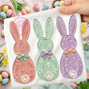 May include: Three decorative Easter bunny figurines in pastel colors: pink, mint green, and lavender. Each bunny has a bow, floral accents, and gold glitter details. The bunnies are displayed on a white surface, with Easter eggs and flowers in the background.