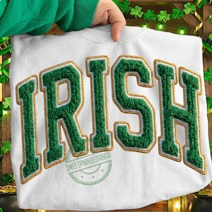Op de afbeelding: Witte sweatshirt met het woord "IRISH" in grote, groene, getextureerde letters met gouden rand. De sweatshirt wordt door een persoon omhoog gehouden. Een feestelijk ontwerp voor St. Patrick's Day.