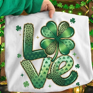 May include: White sweatshirt with a St. Patrick's Day design. The word "LOVE" is spelled out with green leopard print letters and a large green shamrock. Gold glitter outlines the letters and shamrock.