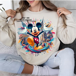 May include: A cream-colored sweatshirt with a colorful graphic print of Mickey Mouse standing in front of a cityscape. The graphic features a colorful swirl of paint and splatters.