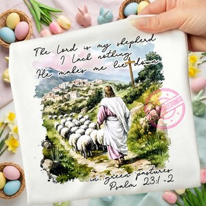 May include: A white t-shirt featuring a watercolor illustration of Jesus leading a flock of sheep. The image includes text from Psalm 23:1-2, with a cross and a village in the background. The design is surrounded by Easter decorations.