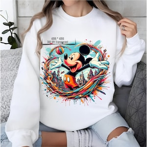 May include: White sweatshirt with a colorful graphic print of Mickey Mouse playing with a ball in front of a castle. The graphic is surrounded by colorful paint splatters.