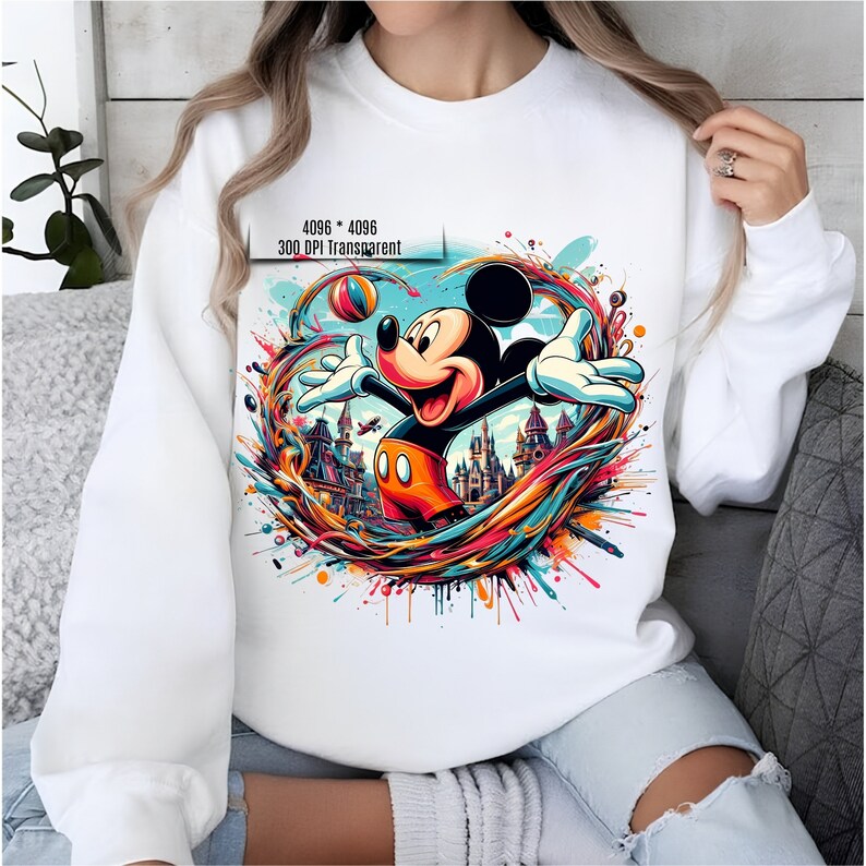 May include: White sweatshirt with a colorful graphic print of Mickey Mouse playing with a ball in front of a castle. The graphic is surrounded by colorful paint splatters.