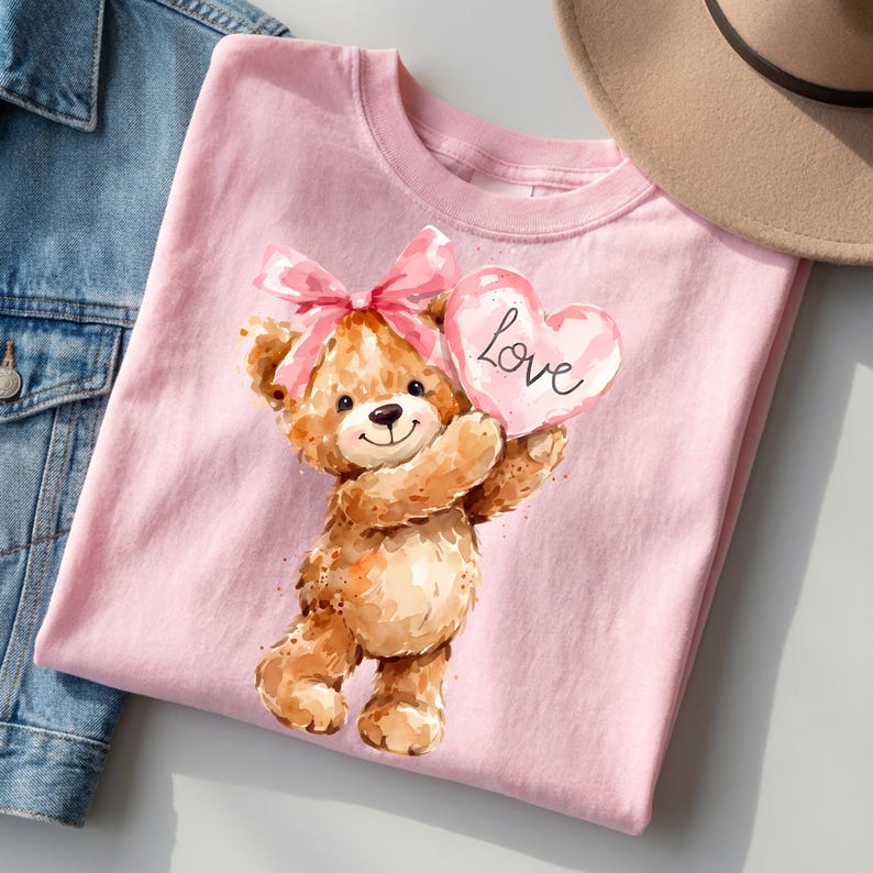 May include: Pink t-shirt featuring a watercolor illustration of a teddy bear holding a heart-shaped balloon with the word "Love". The bear has a pink bow and is smiling. A denim jacket and a tan hat are in the background.