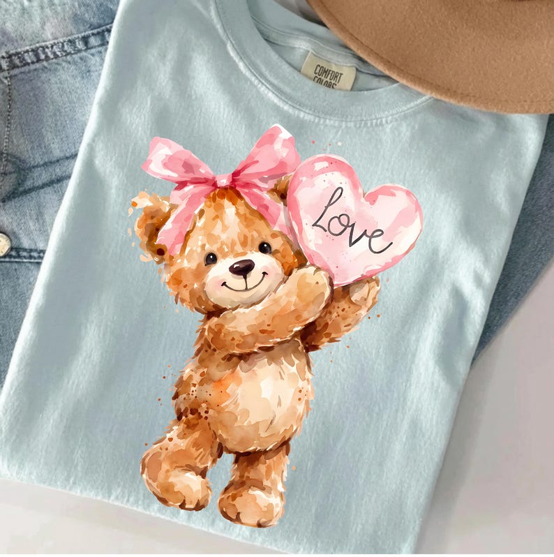 May include: Light blue t-shirt featuring a watercolor illustration of a teddy bear holding a pink heart-shaped candy with the word "Love" written on it. The teddy bear has a pink bow on its head.