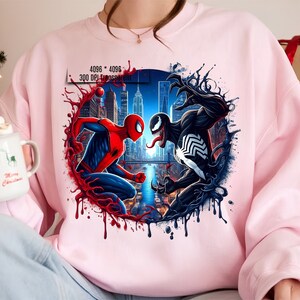 May include: A pink sweatshirt with a graphic print of Spider-Man and Venom fighting in a city setting. The graphic is surrounded by red and black dripping paint.