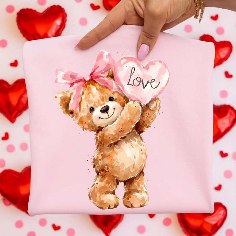 May include: A pink sweatshirt featuring a watercolor illustration of a teddy bear holding a heart-shaped balloon with the word "Love" on it. The bear has a pink bow and is surrounded by red heart-shaped balloons.