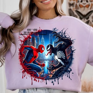 May include: A purple sweatshirt with a graphic print of Spider-Man and Venom fighting in a city setting. The graphic is surrounded by red and black splatters.