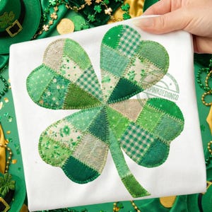 Può includere: Una maglietta bianca con un grande design a patchwork a forma di quadrifoglio. Il quadrifoglio è composto da vari tessuti verdi con diversi motivi, tra cui floreali, a quadri e tinta unita. Il disegno è tenuto in mano, con accessori a tema San Patrizio sullo sfondo.