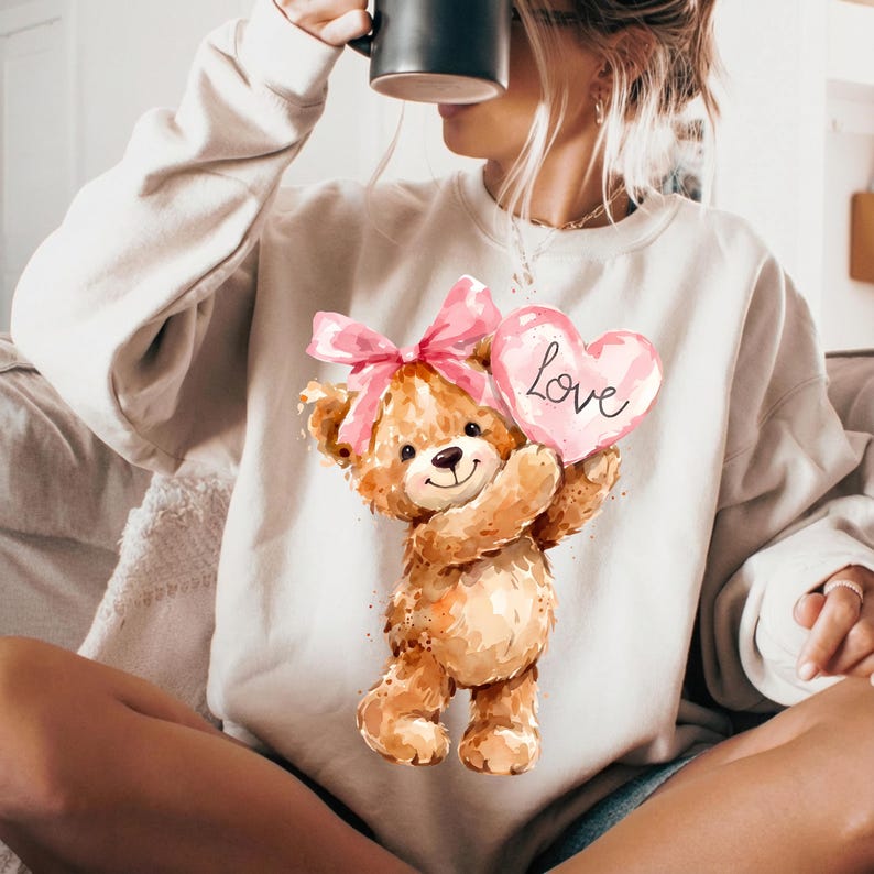 May include: A beige sweatshirt featuring a watercolor illustration of a teddy bear holding a pink heart with the word "Love" on it. The teddy bear has a pink bow and is holding the heart. The sweatshirt is a neutral color.