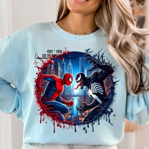 May include: A light blue sweatshirt with a graphic print of Spider-Man and Venom fighting in a city setting. The graphic is surrounded by red and black dripping paint.