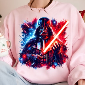 May include: A pink sweatshirt with a graphic print of Darth Vader holding a red lightsaber. The background is a galaxy with blue and red stars.